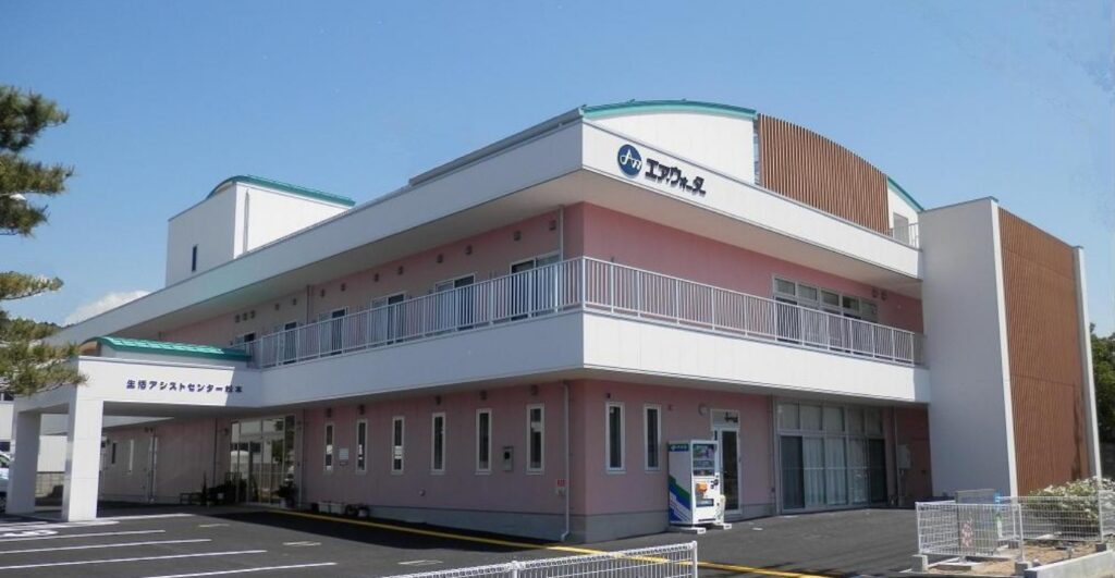 介護施設の建物外観。白とピンク色の壁、緑色の屋根が特徴の2階建て施設